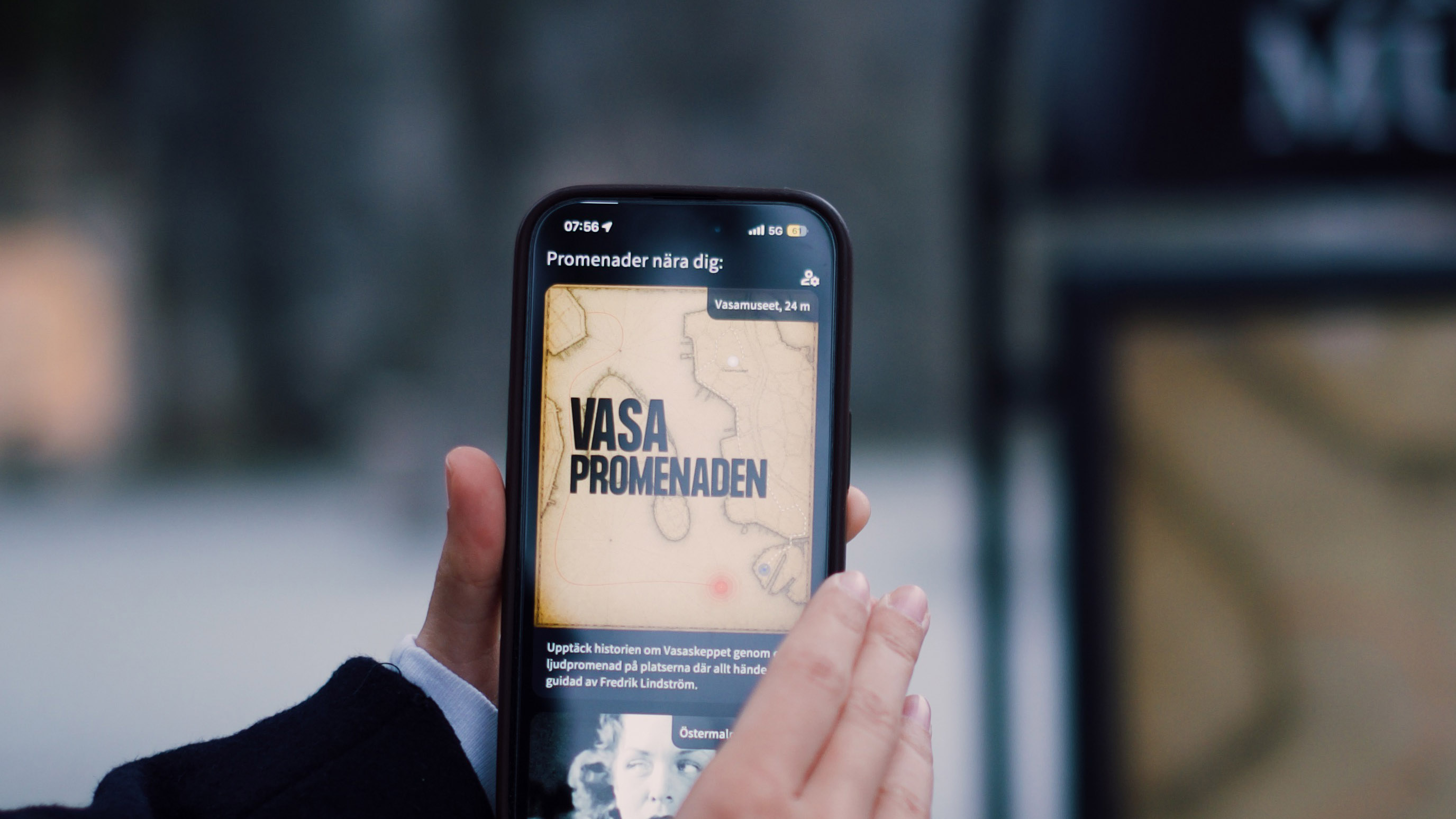 Foto av hand som håller i en telefon med appen Vasapromenaden öppen.