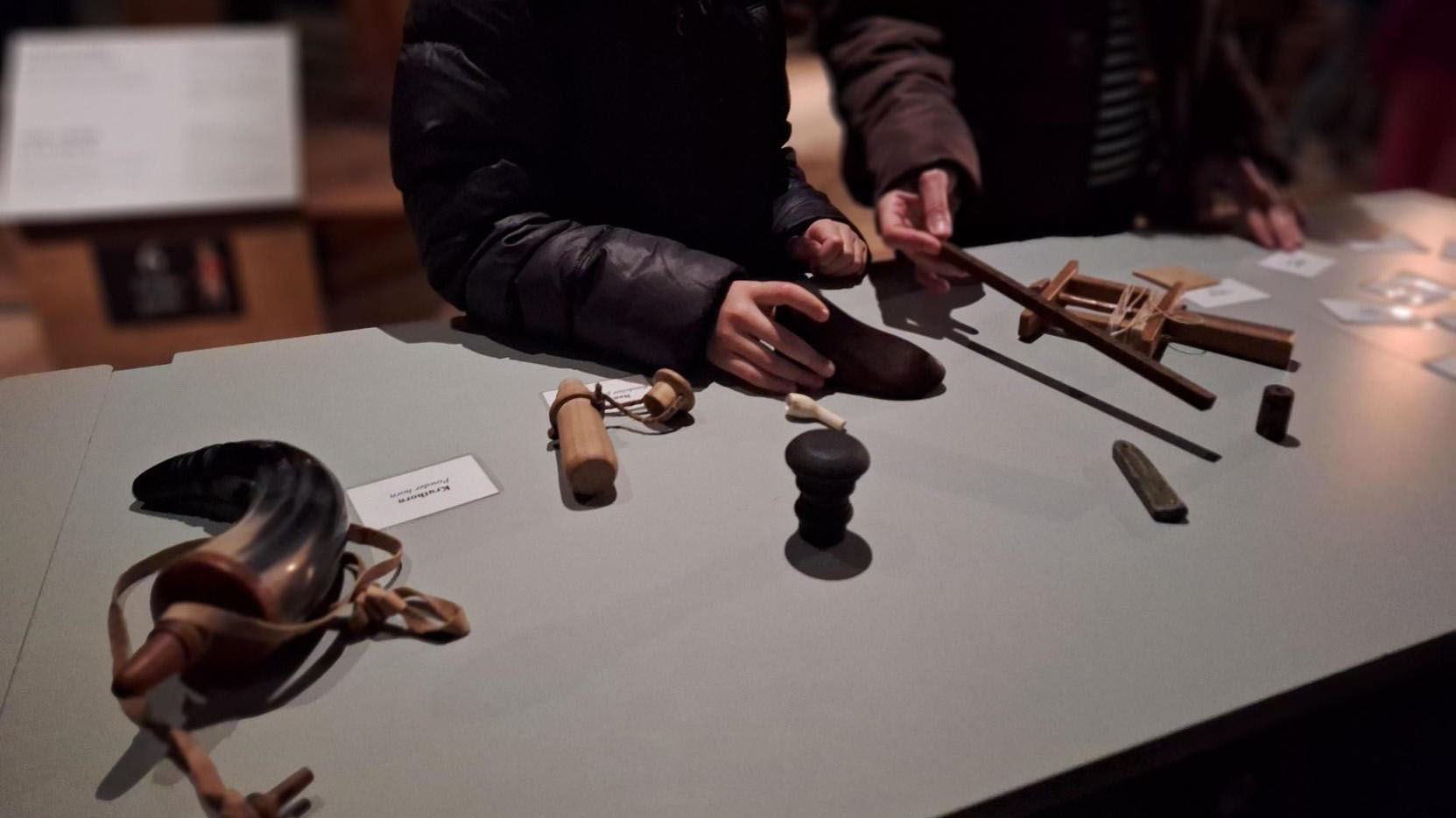 Ett bord med föremål i olika former, person klädd i tjock jacka sitter vid bordet. 