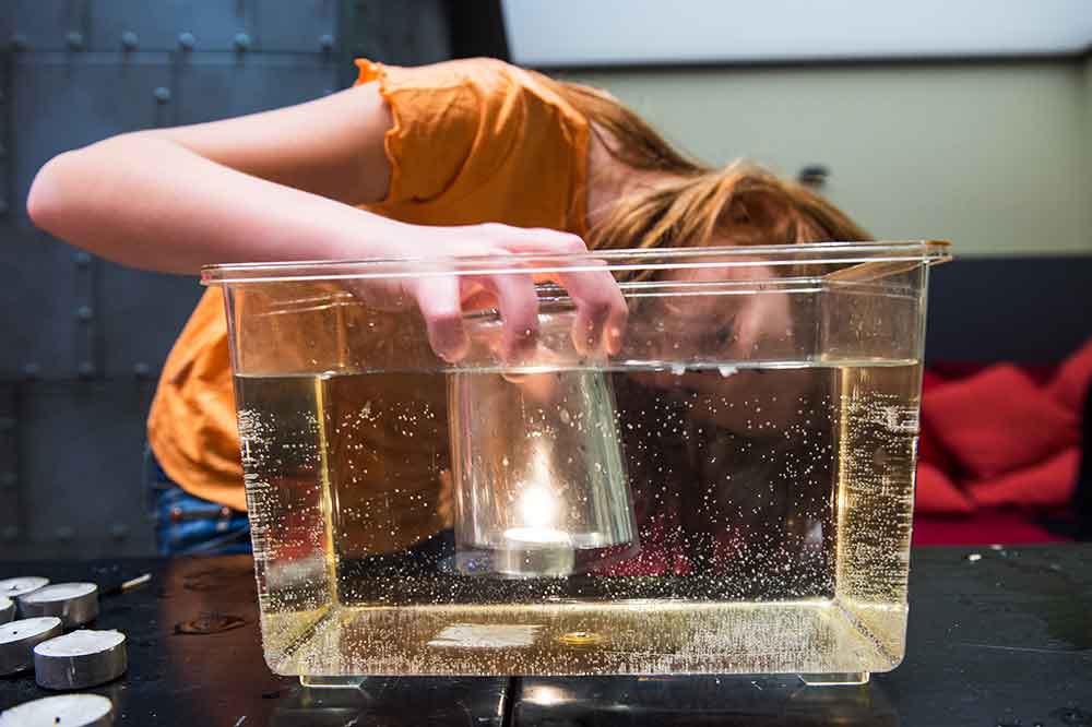 Flicka i gul tröja håller handen över genomskinligt glasi en vattenbehållare. Ett tänt värmeljus lyser i glaset. 