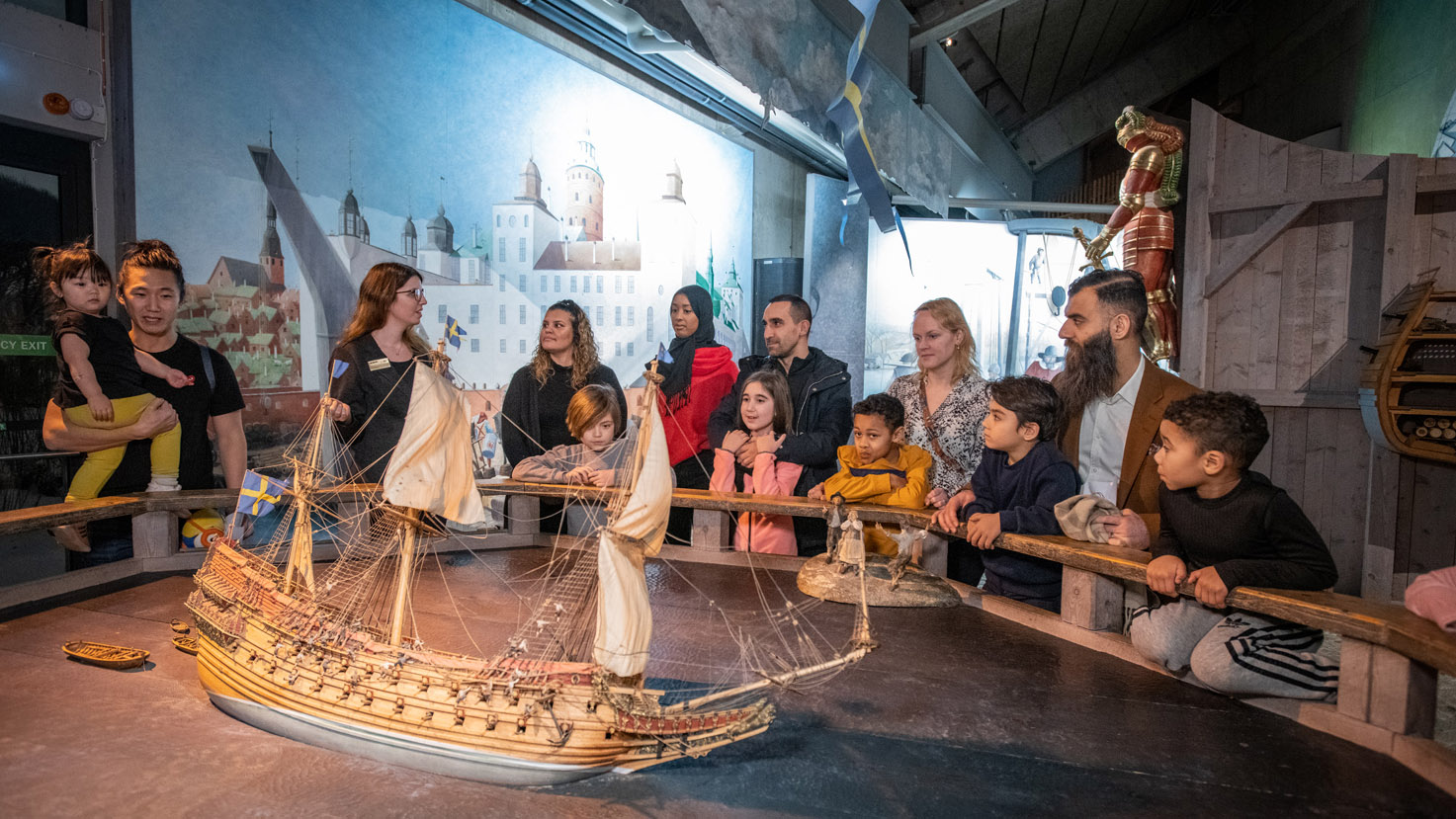 En grupp människor i olika åldrar och etnicitet lyssnar på en guide vid en modell av skeppet Vasa.