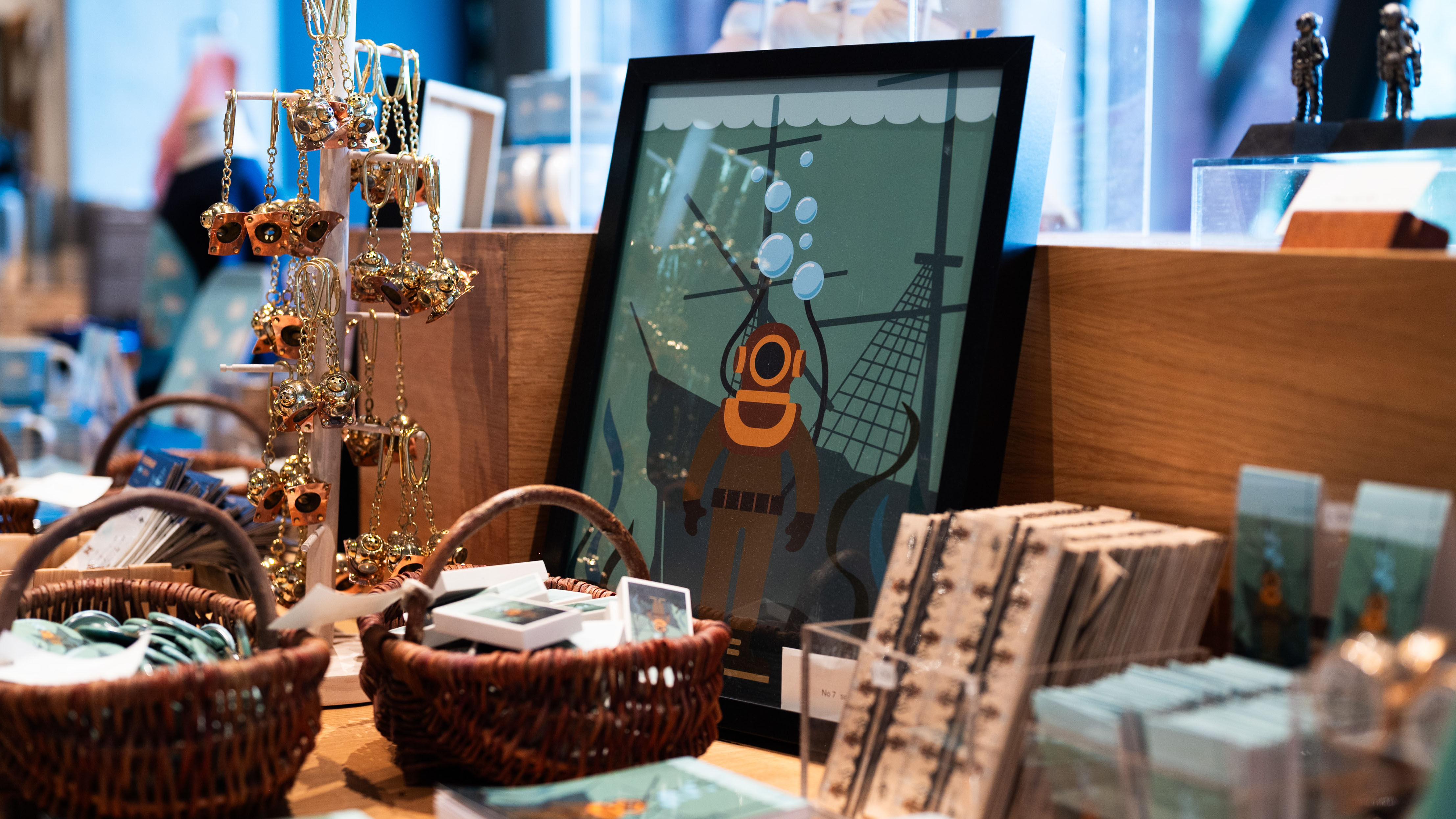 Närbild av museibutikens hylla med en inramad illustration av en dykare och ett skepp, omgiven av korgar och smycken i mässingstoner.