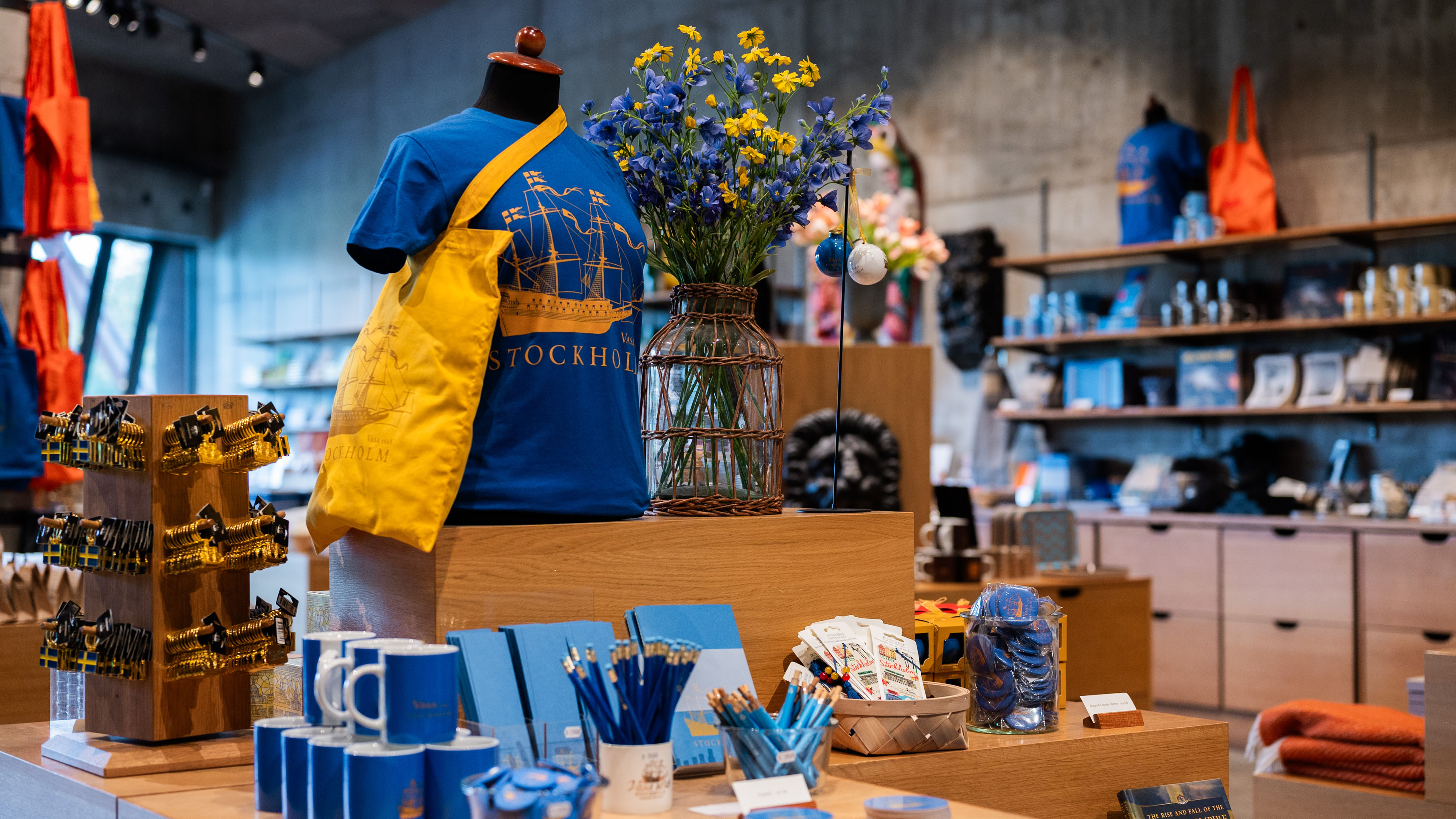 Butiksdisplay med blå och gula produkter, inklusive en t-shirt med Stockholmsmotiv och en tygpåse, dekorerad med blommor i vas.