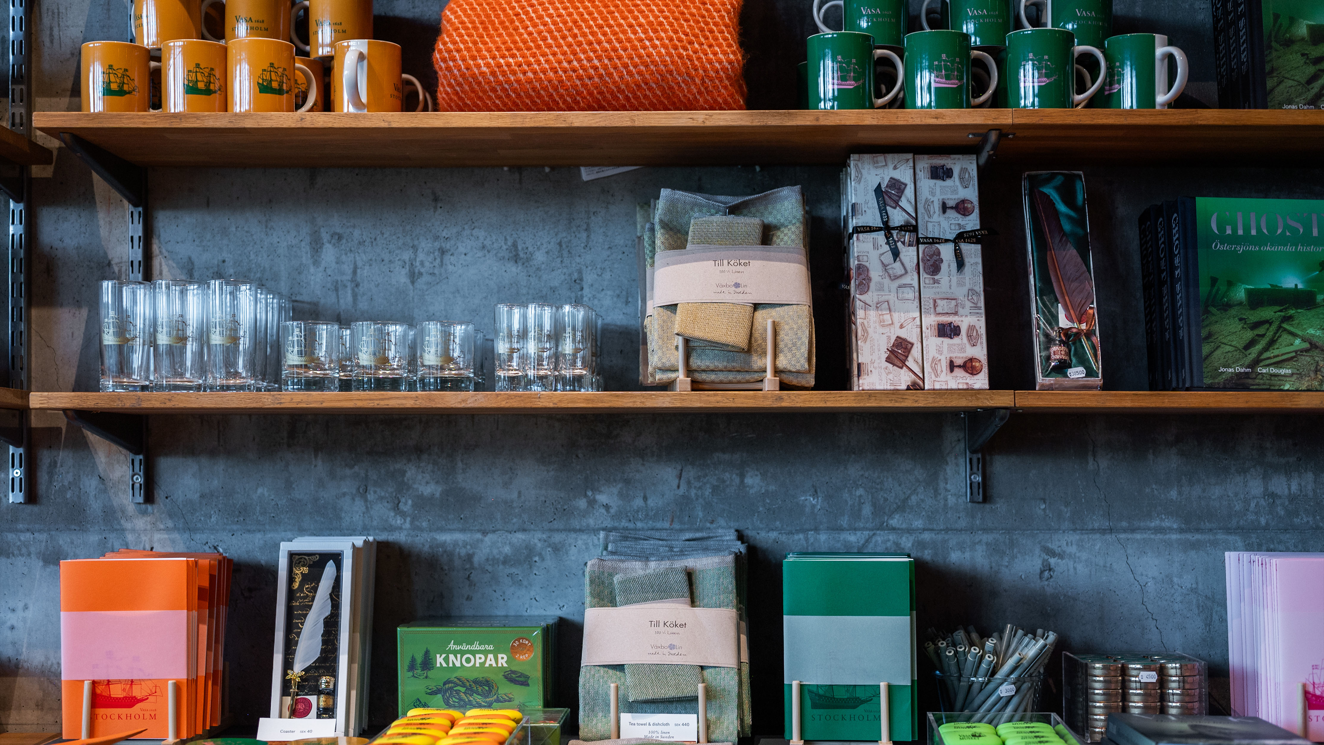 Hylla i museibutik med kopparmuggar, glas, handdukar och böcker, arrangerade mot en betongvägg.