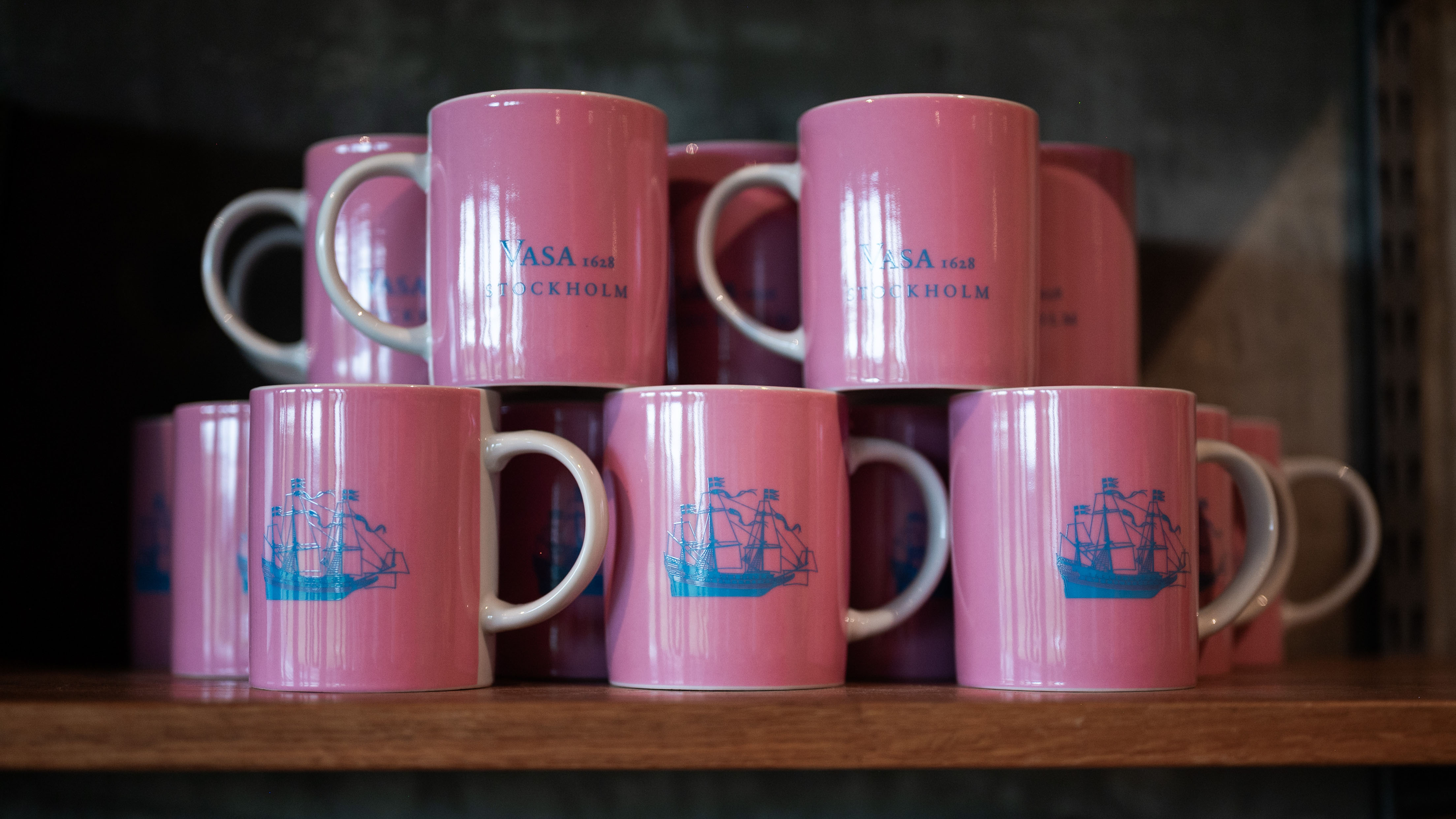 Stapel av rosa muggar med blå skeppsmotiv och texten ”Vasa 1628 Stockholm” på en trähylla i museibutiken.