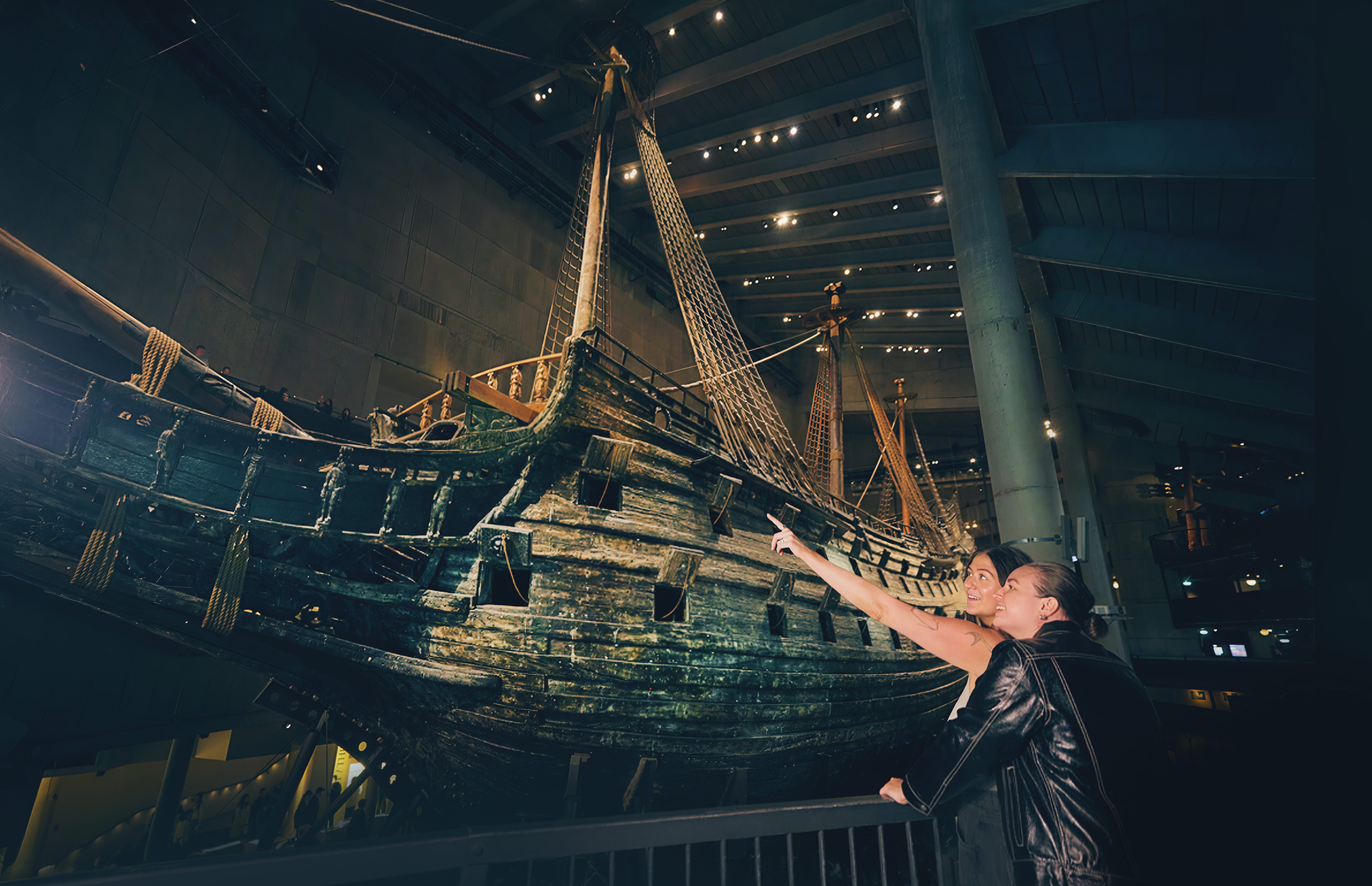 Foto innifrån Vasamuseet, två kvinnor står tätt intil varandra och tittar på skeppet