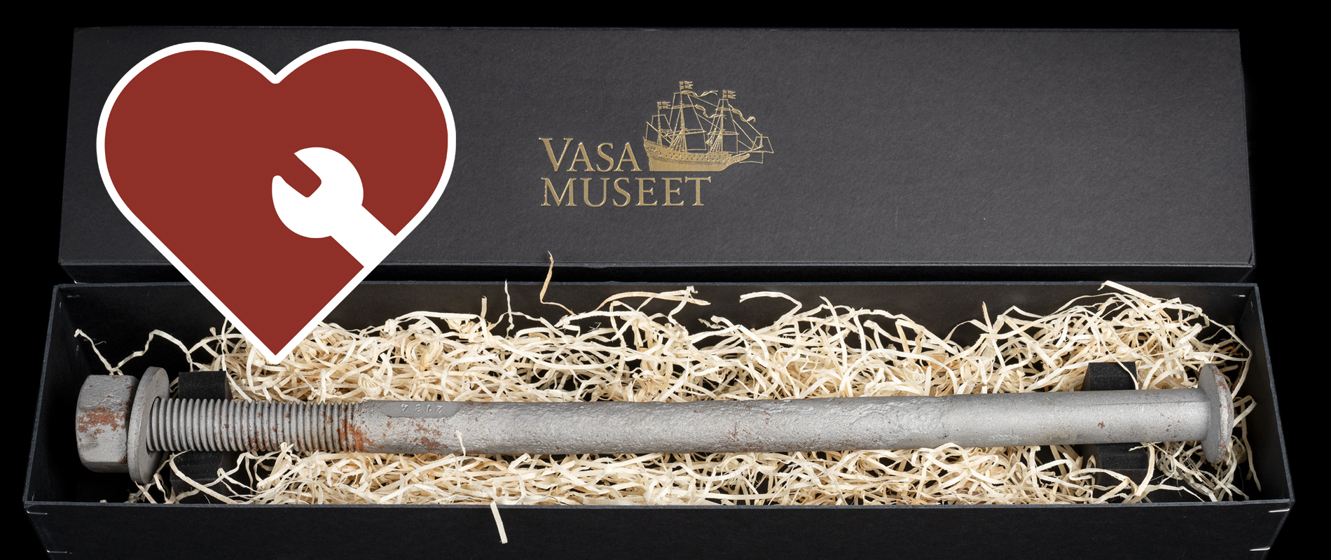 Foto av bult från skeppet Vasa i mörk låda med träull. Ett rött hjärta med vit kant och vit skiftnyckel syns ovanpå bilden. 