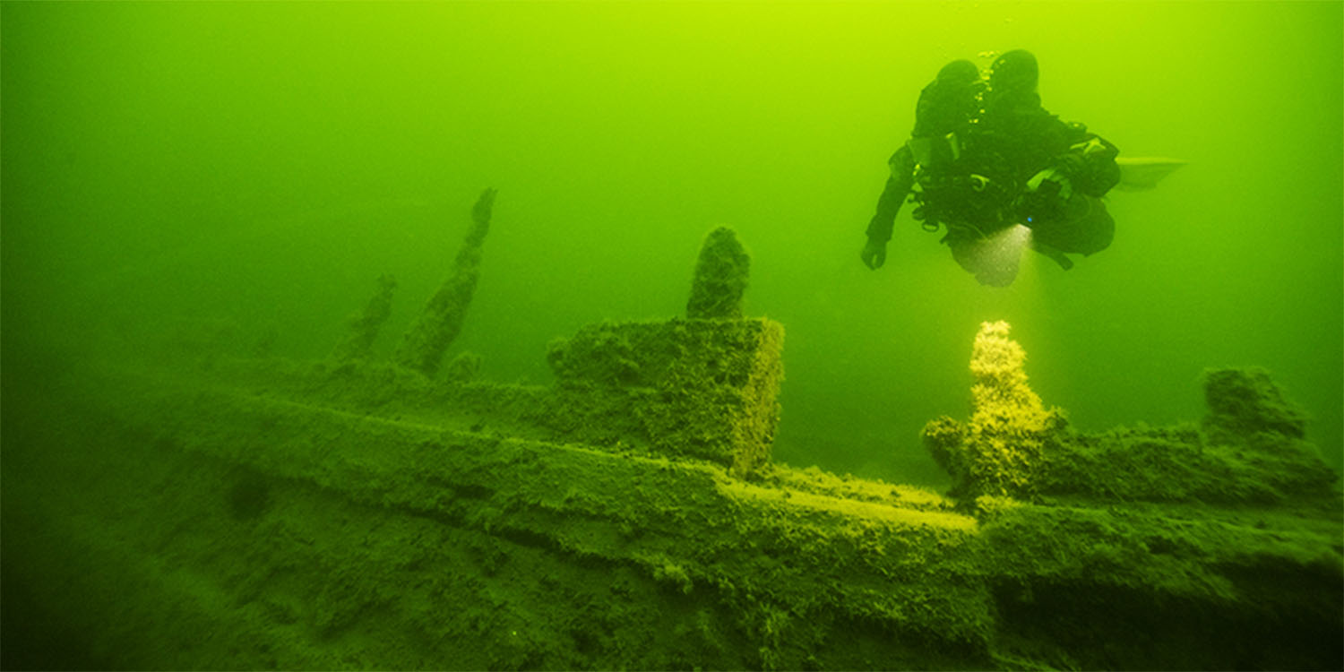 Gröntonad undervattensbild som visar en dykare ovandör ett upprättstående vrak övervuxet med alger.