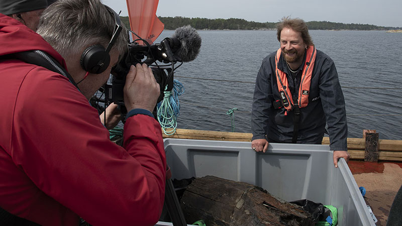 Pressuppbåd samlat runt tunna som ligger i grå låda, marinarkeolog JIm Hansson visar upp. 