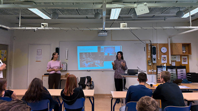 Två av Vasamuseets anstäldla står längst fram i ett klassrum, med en powerpoint-presentation i bakgrunden. Elevers nackar och bakhuvuden syns. 