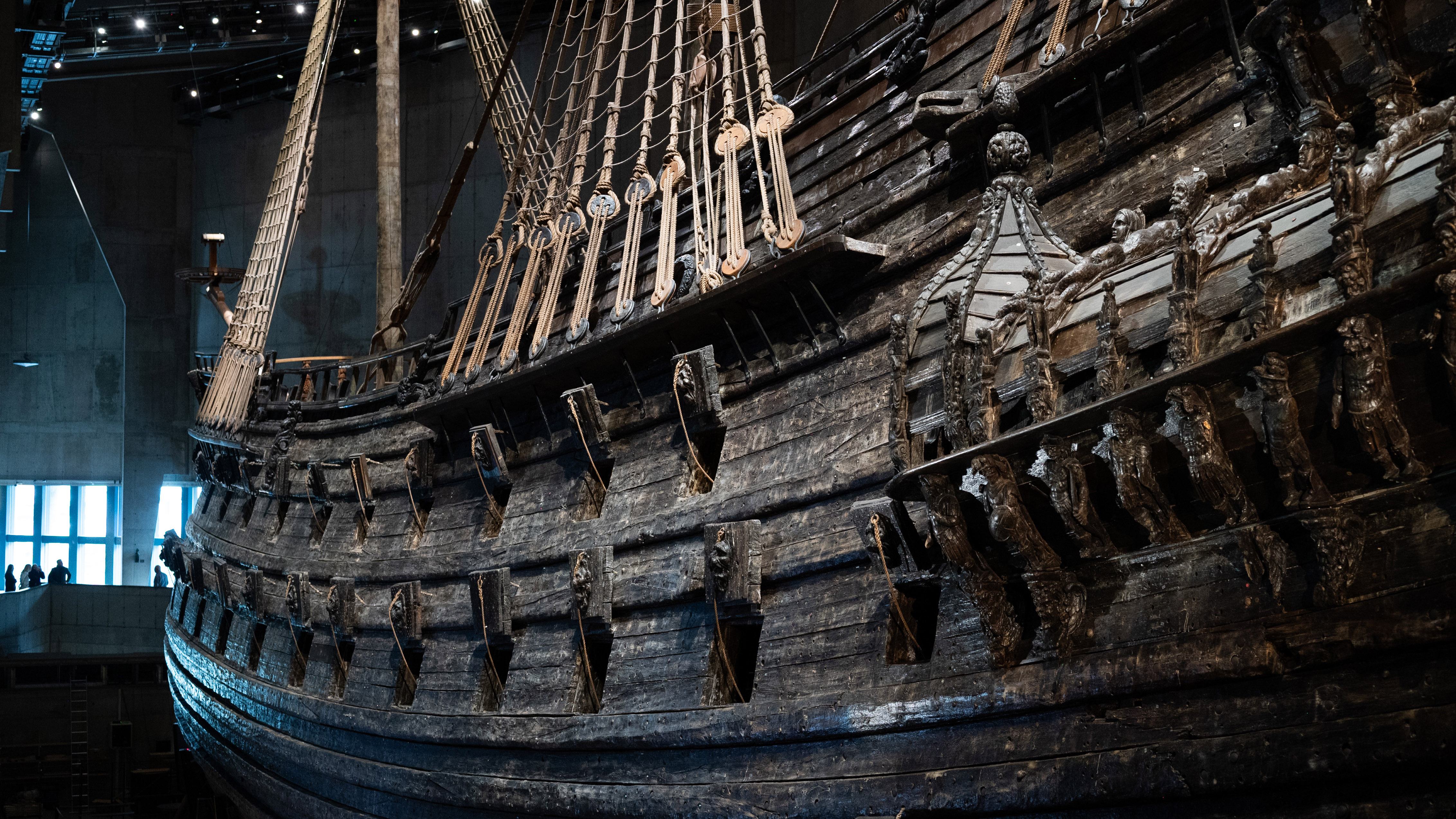 Foto av skeppet Vasa inne i museet sett från ena sidan. Kanonluckorna är öppna,  byggnaden dunkel. 
