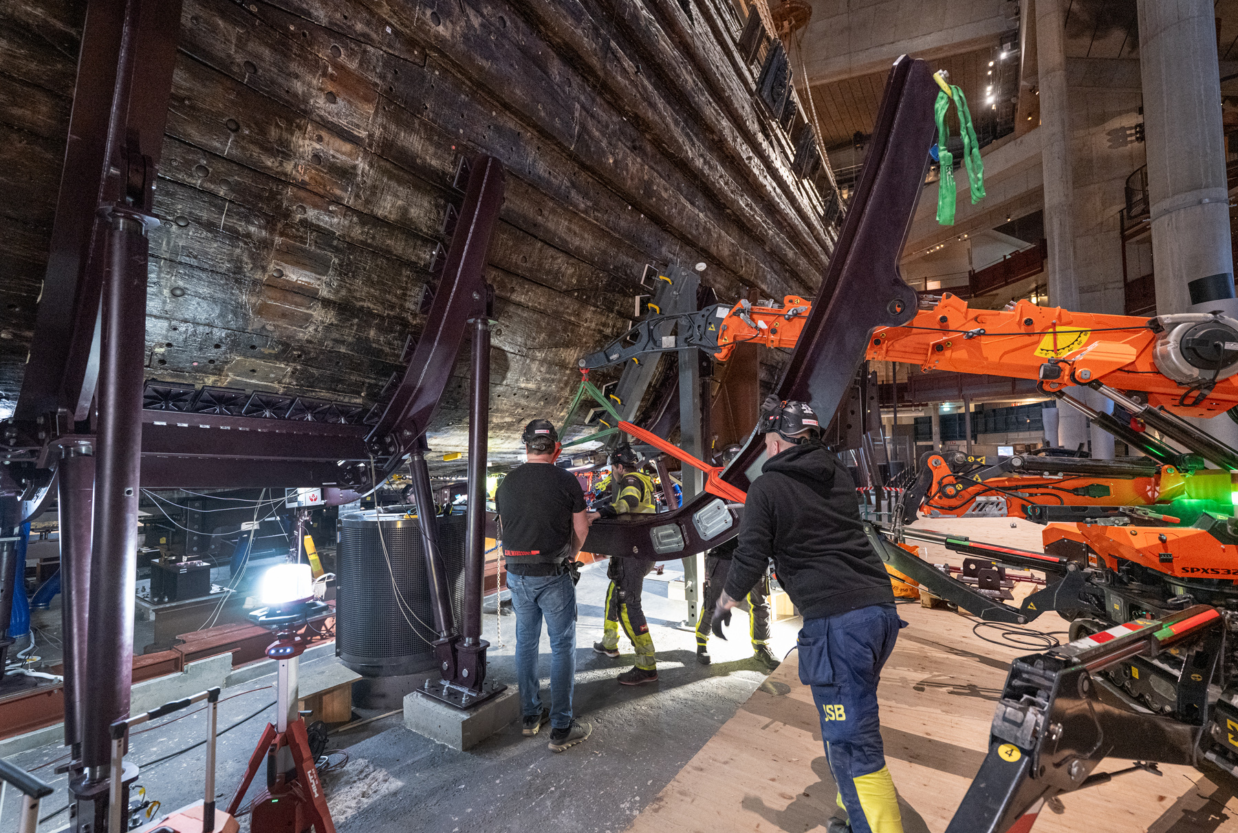 Tre personer i arbetskläder och hjälmar invud skeppet Vasas undersida med stöttor och orange kran.